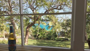 A bottle of Kalik beer on a wooden table by a large window with a view of a garden and a blue sky. - Beautiful Hibiscus House (Governor's Harbour)