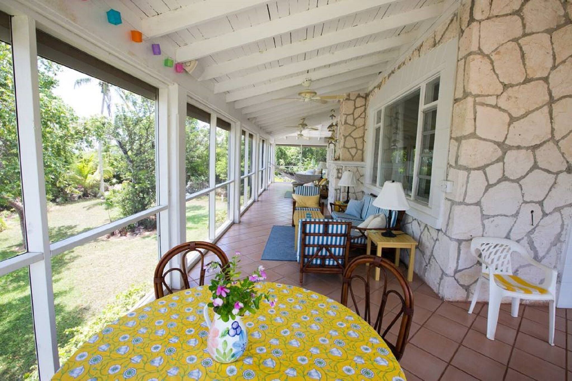 A covered patio with a stone wall, a dining table with a yellow floral tablecloth, and a hammock.
