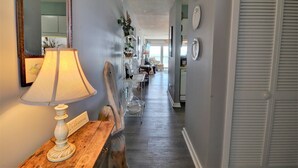 A hallway with a wooden bench, a lamp, and a mirror on the left. There are shelves with decorative items and a view of a dining area in the background. - Oceanfront 2-bedroom Queen's Grant Condo (Topsail Beach)