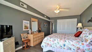 A bedroom with a bed, a dresser, a television, a ceiling fan, and a window with closed blinds. - Oceanfront 2-bedroom Queen's Grant Condo (Topsail Beach)
