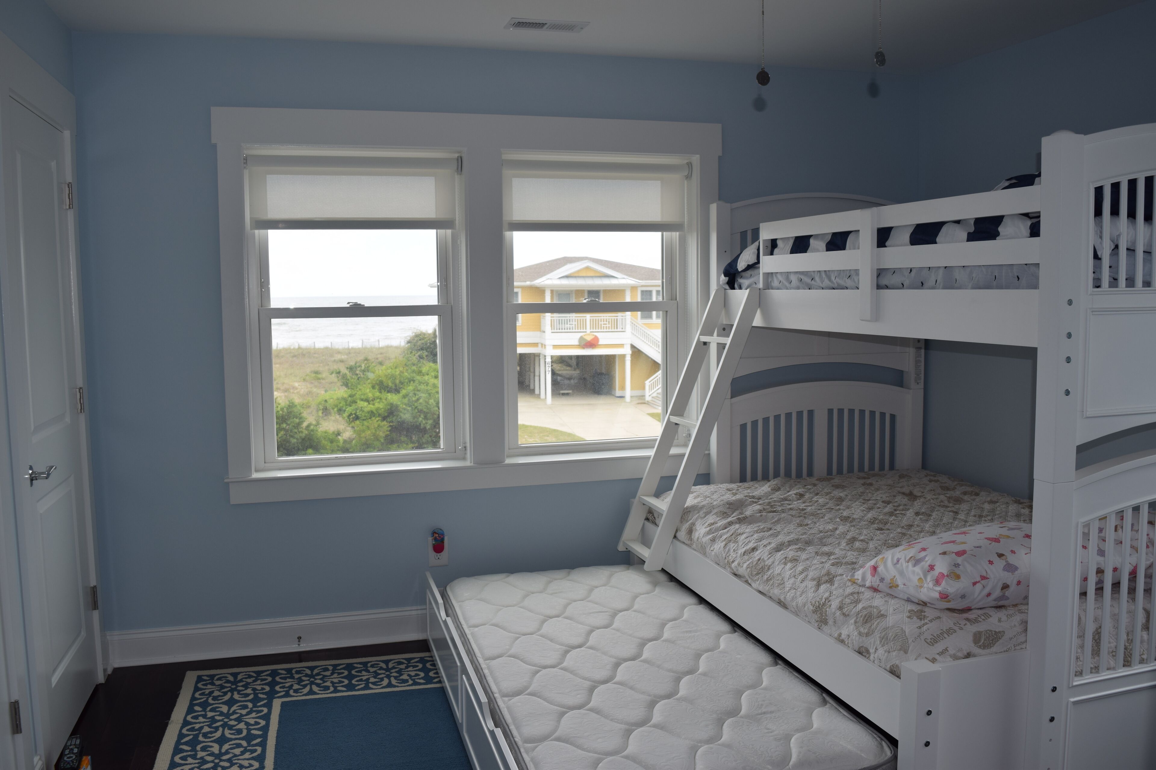 Une chambre avec un lit superposé, une fenêtre donnant sur une maison et de la verdure, et une porte.