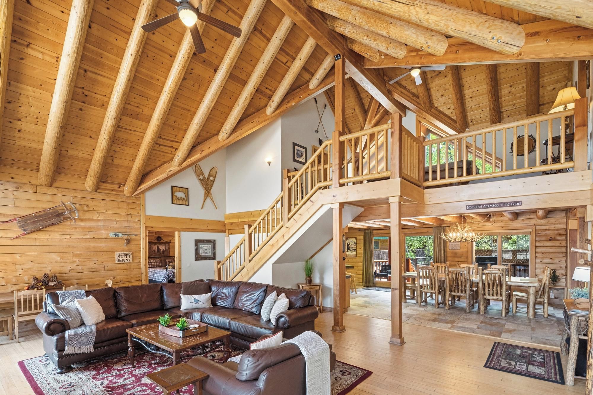 Un intérieur de cabine spacieux, avec un plafond haut, des poutres en bois, un espace de vie central comprenant un canapé et une table basse, un escalier menant à un niveau supérieur et un coin repas avec des meubles en bois.