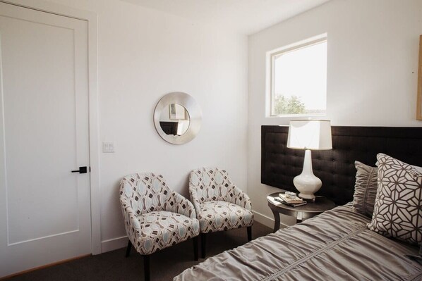 A bedroom with a bed, two patterned chairs, a round mirror, a nightstand with a lamp, and a window providing natural light.