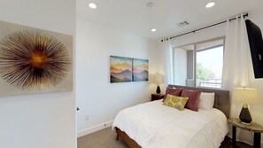 A bedroom with a bed, two wall-mounted lamps, a window with curtains, and two paintings on the wall.