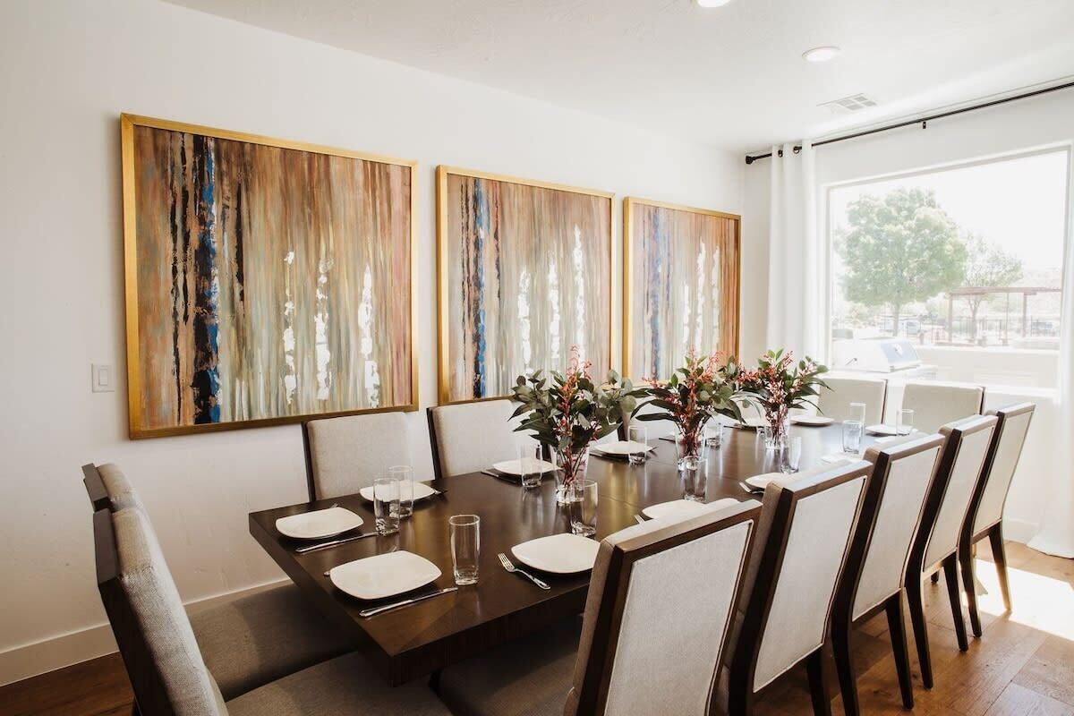 A dining area with a rectangular table set for six, flanked by two large abstract paintings, and a window providing a view of the outdoors.