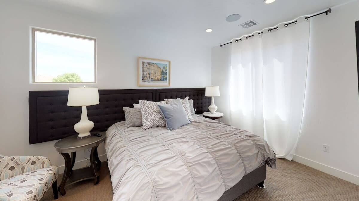 A bedroom with a large bed, two bedside tables, a nightstand, a window with curtains, and a wall-mounted artwork.