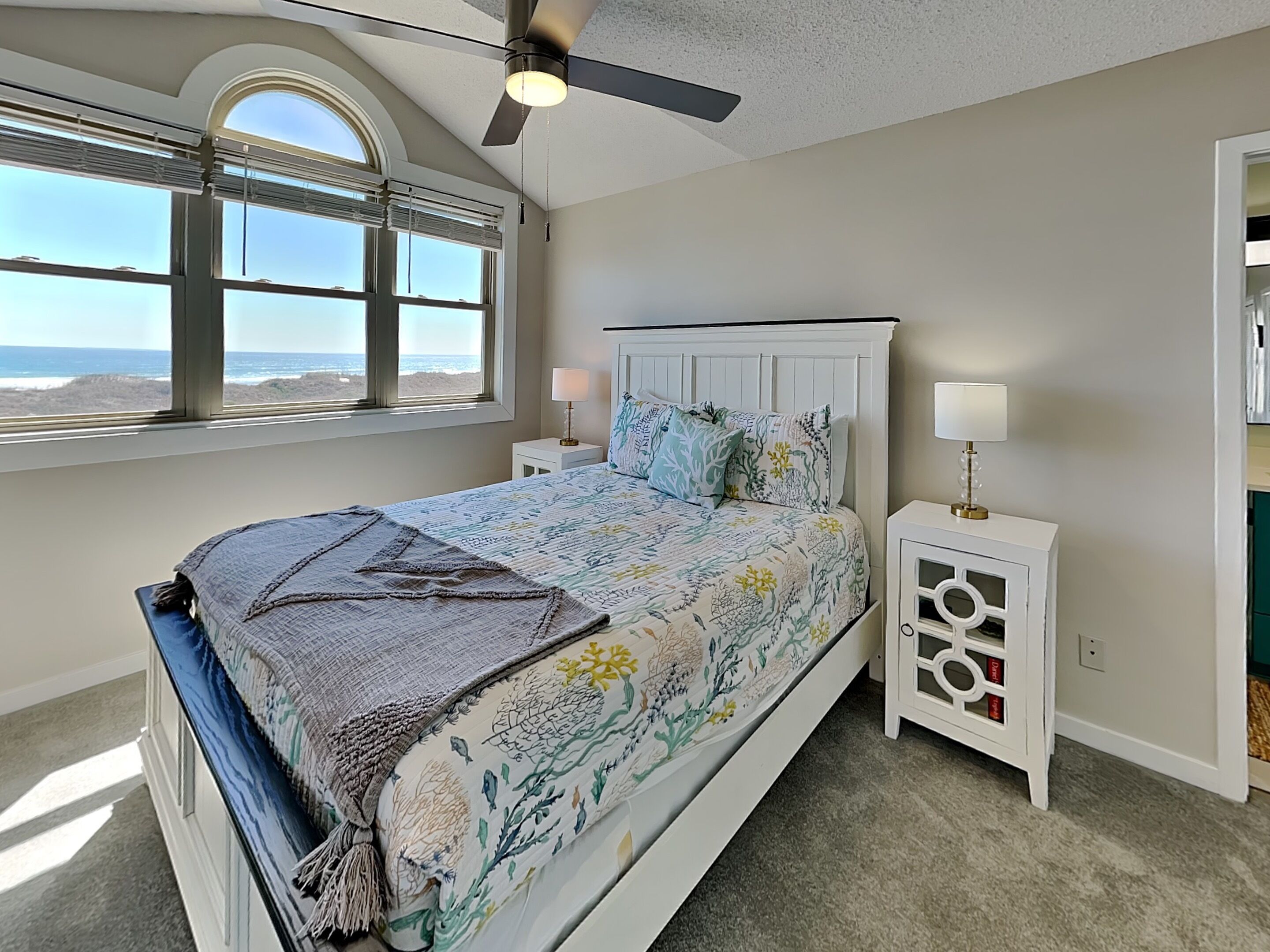 A bedroom with a bed, a ceiling fan, a nightstand with a lamp, a window with a view of the ocean, and a built-in cabinet.
