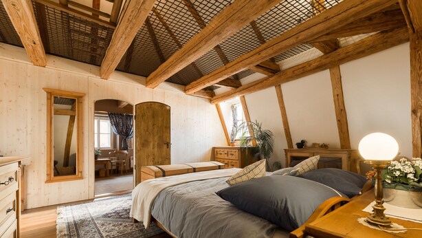 A bedroom with a wooden ceiling, a bed with grey and blue bedding, a wooden dresser, and a nightstand with a lamp. - Doppler's Apartment fully A/C (Prague)