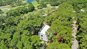 A large, white, two-story house surrounded by dense green trees, with a golf course and ocean view in the background. - Unique Home in Dauphin Island's Maritime Forest - Stunning Home and Water Views (Dauphin Island)