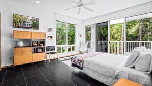 A modern bedroom with a large bed, a ceiling fan, a wooden cabinet, and large windows offering a view of trees. - Unique Home in Dauphin Island's Maritime Forest - Stunning Home and Water Views (Dauphin Island)