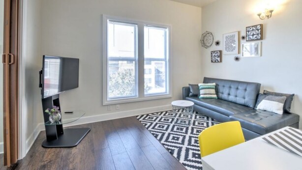 Ein modernes Wohnzimmer mit grauem Sofa, einem kleinen runden Couchtisch und einem Fernseher auf einem Lowboard. Große Fenster mit Jalousien, ein gemustertes Teppich und Wandbilder sind ebenfalls zu sehen.