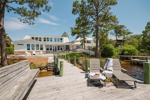 Um deck de madeira com cadeiras de descanso e uma pequena piscina, cercado por árvores e um edifício com grandes janelas.