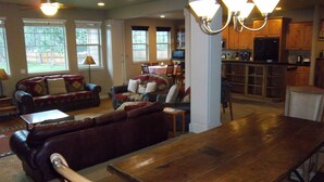 A spacious living room with a large wooden dining table, a dark leather sofa set, and a kitchen area with wooden cabinets and a built-in oven. - Skyline Lodge, Gorgeous Setting, Walk to Downtown! (Bend)