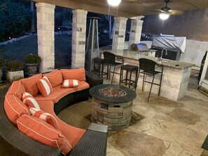 An outdoor patio with a stone fire pit, a curved sofa with orange cushions, and a bar area with stools. - Peaceful, Super Clean Central SA Home (San Antonio)
