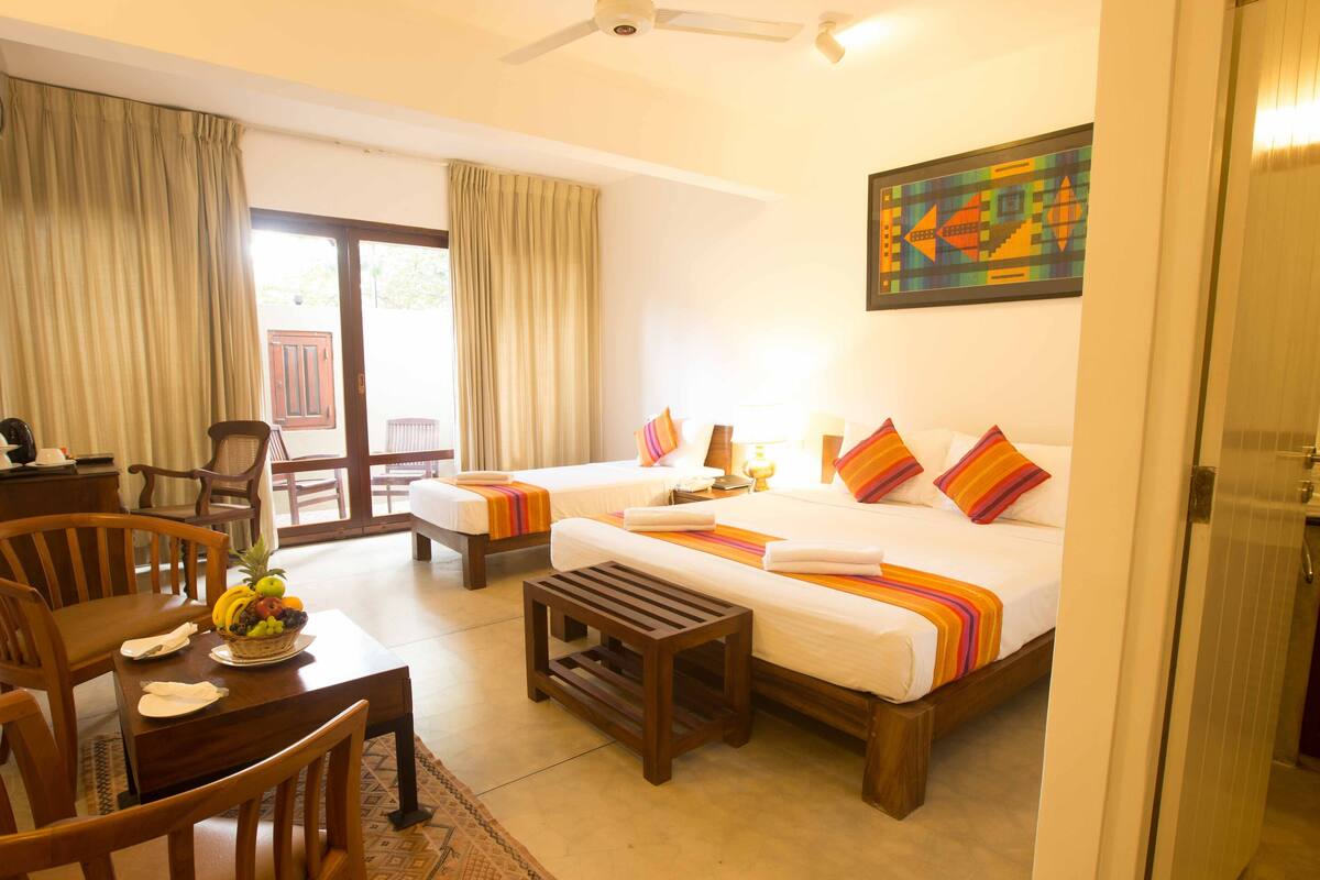 A hotel room with two beds, a dining table with chairs, a small wooden table, and a balcony with chairs.