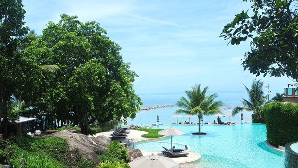 Piscina de resort com vista para o oceano, rodeada por vegetação exuberante e palmeiras.