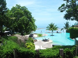 Piscina de resort con vista al océano, rodeada de exuberante vegetación y palmeras.