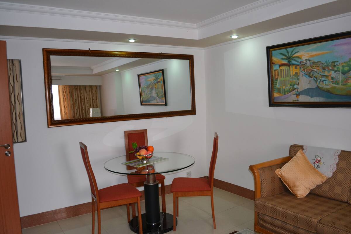 A dining area with a glass table, two chairs, and a sofa with a cushion.