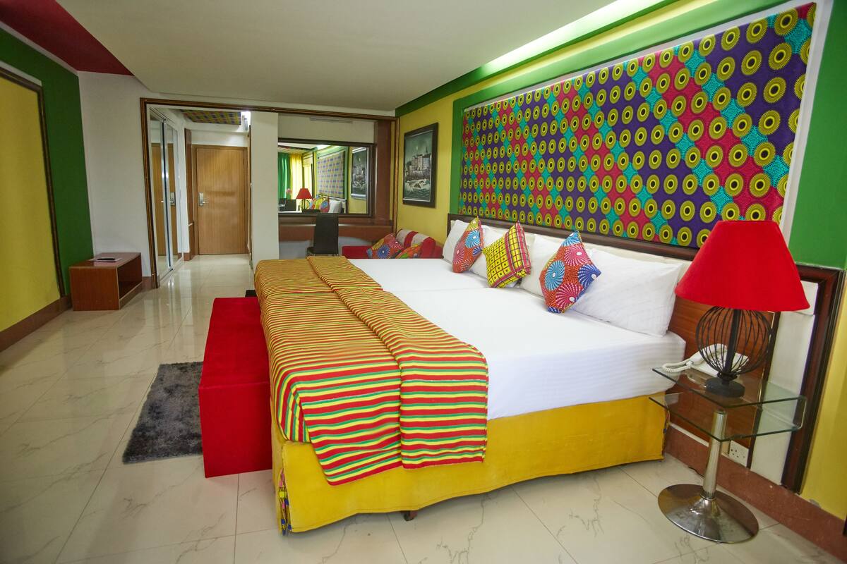 A hotel room with a large bed, a bedside table, a red lamp, and a colorful wall pattern.