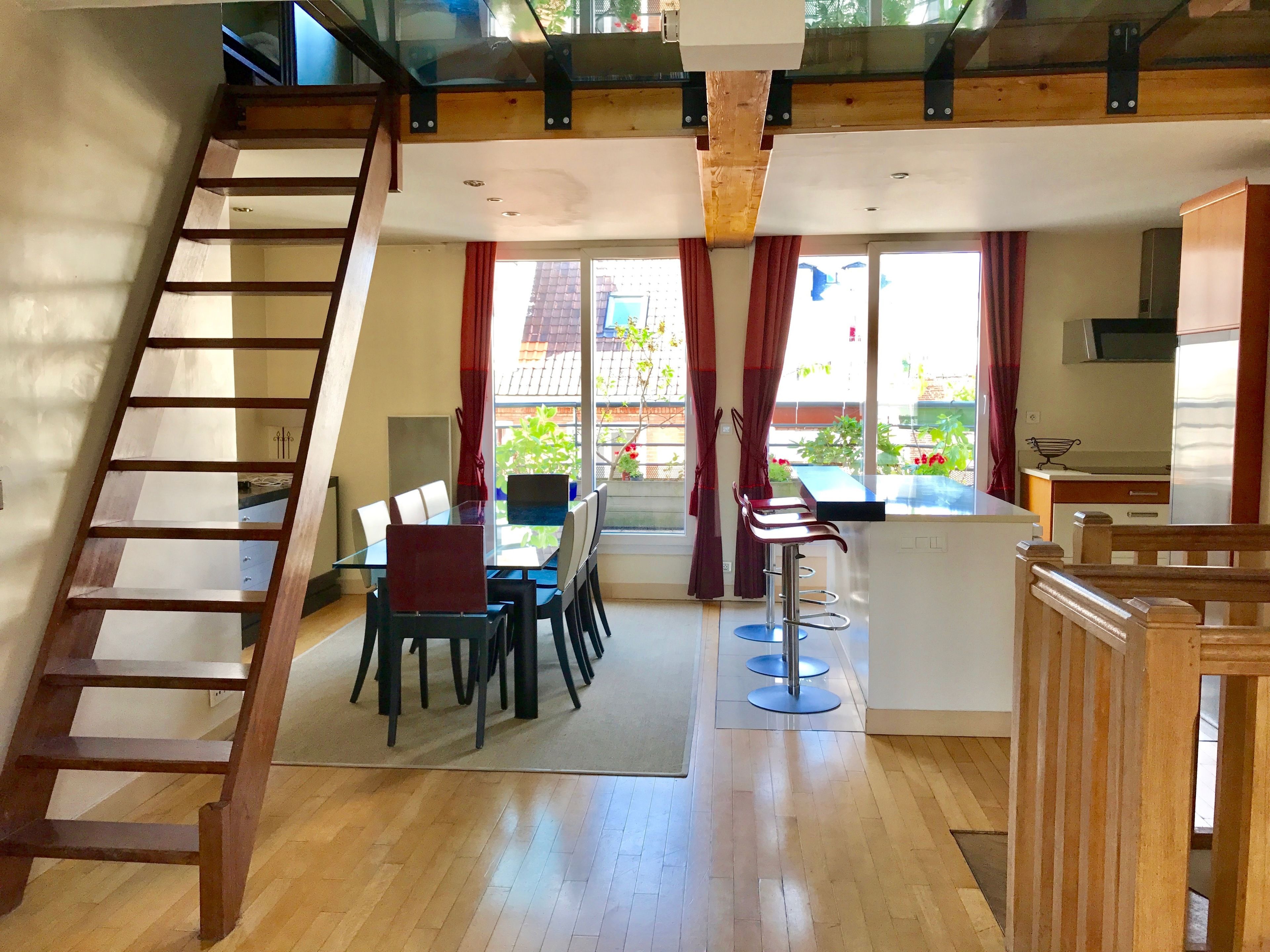 A spacious room with a wooden staircase, a dining area with a glass table, and a kitchen area with a bar counter.