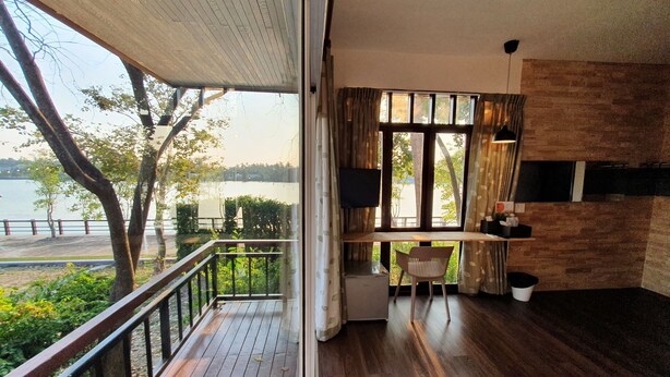 A room with a wooden floor, a desk with a chair, a TV, and a view of a lake. - Baan Tai Had Resort (Samut Songkhram)