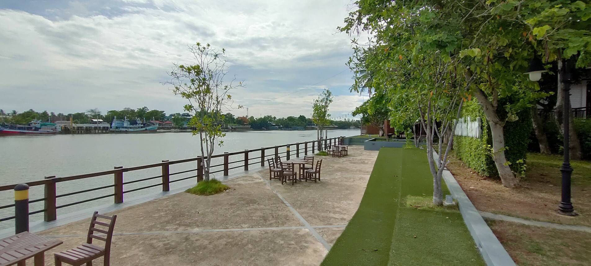 Un paseo fluvial con mesas y sillas, una barandilla y árboles.