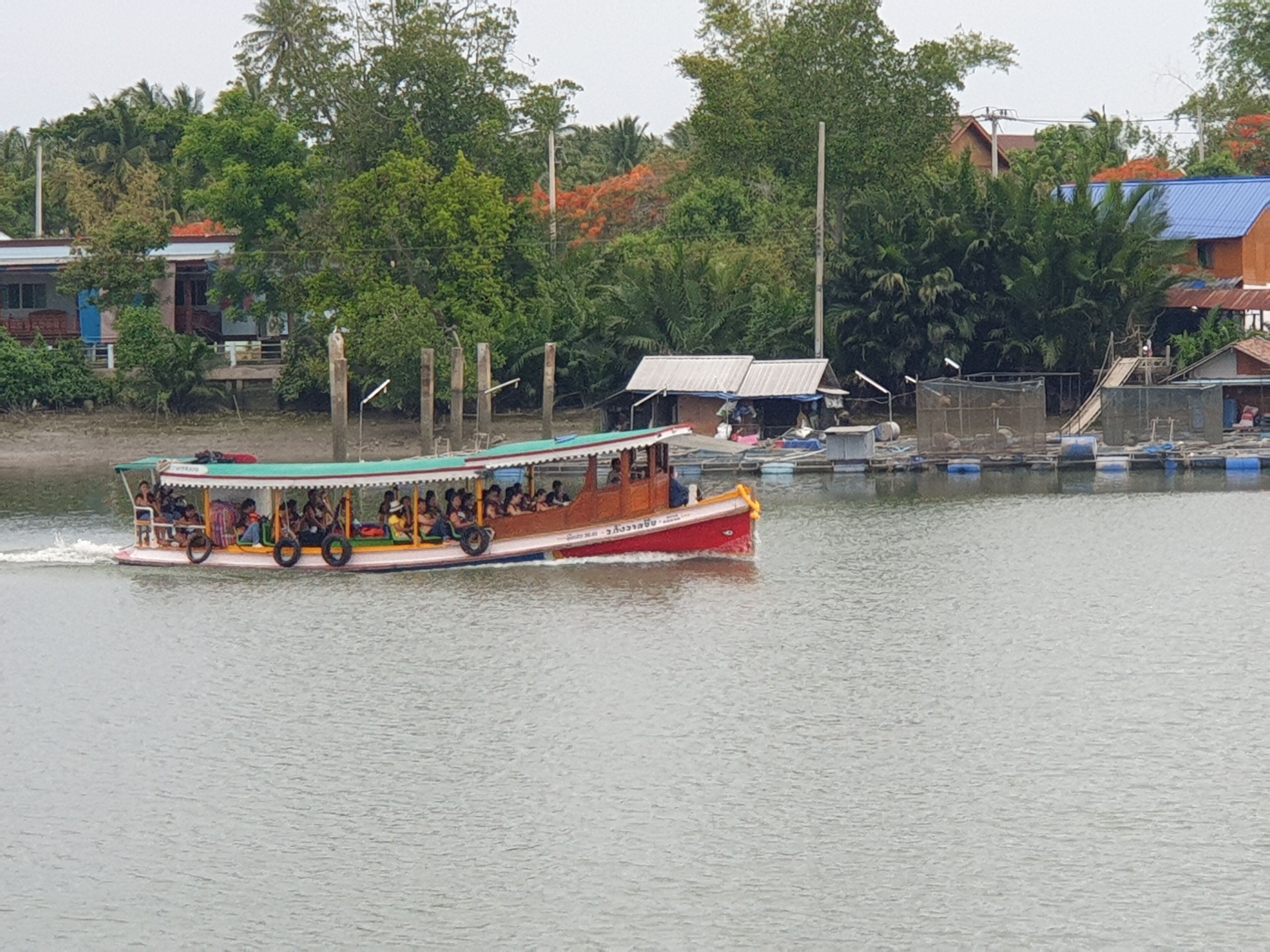 A boat with passengers, moving on a river with buildings and trees in the background.
