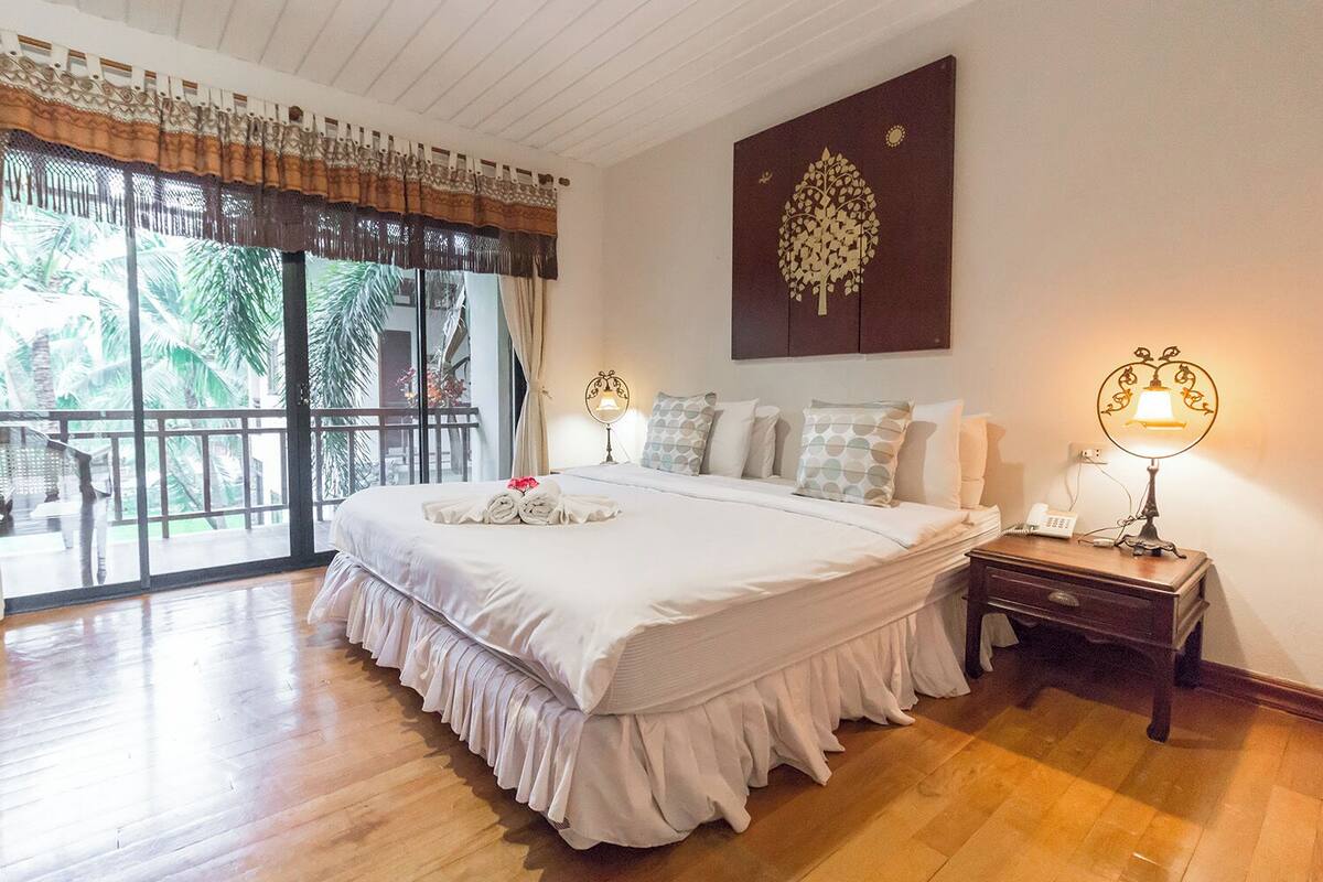 A spacious bedroom with a large bed, a balcony with a view of palm trees, and a wall-mounted artwork.