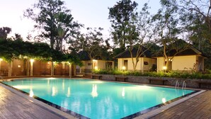 A rectangular swimming pool with a wooden deck surrounding it, set against a backdrop of trees and buildings with warm lighting. - Baan Tai Had Resort (Samut Songkhram)