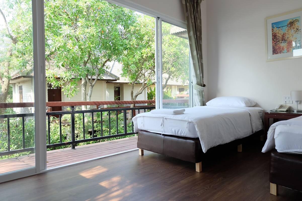 A bedroom with a large window, a bed, a bedside table, and a view of greenery outside.