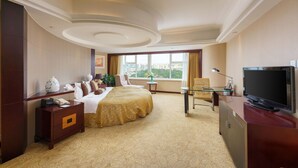 A hotel room with a bed, a desk with a television, a chair, a nightstand, and a window with a city view. - Oasis Grand Hotel (Ürümqi)