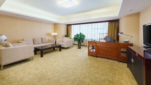 A hotel room with a sofa, a coffee table, a desk with a computer, a television, and a large window with curtains. - Oasis Grand Hotel (Ürümqi)