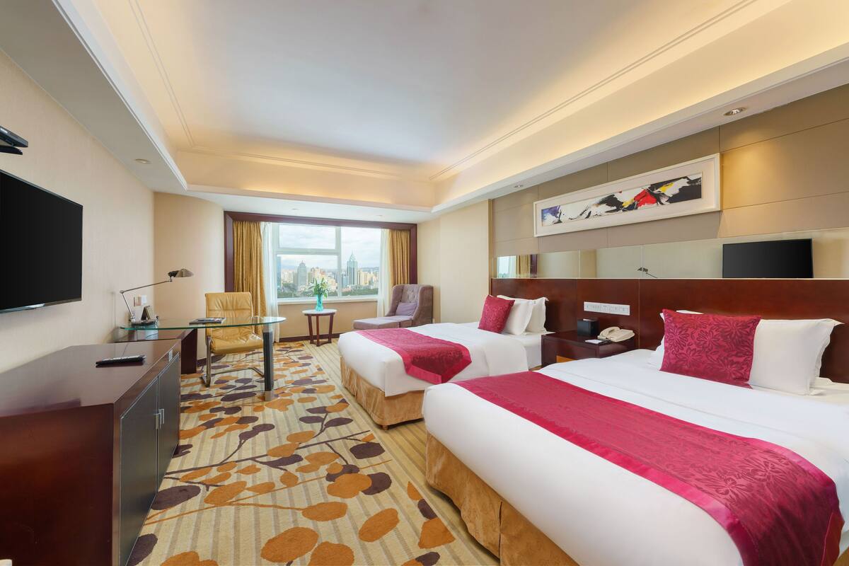 A hotel room with two beds, a desk, a TV, and a window with a city view.