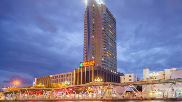 A tall, modern hotel with a prominent vertical light display on top, a covered pedestrian bridge, and a river in the foreground. - Oasis Grand Hotel (Ürümqi)