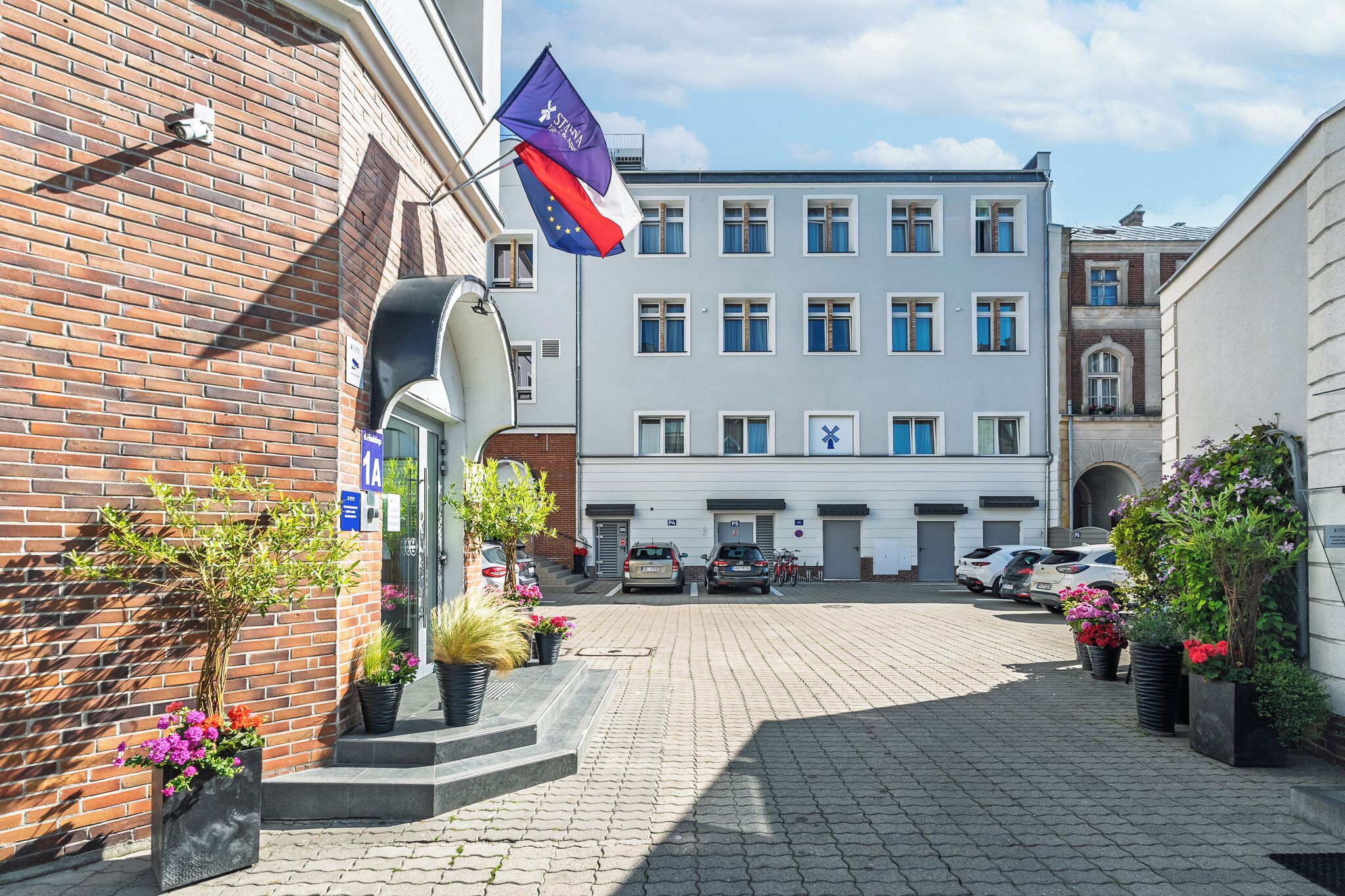 A building with a brick facade, a flag, and a paved courtyard with parked cars.