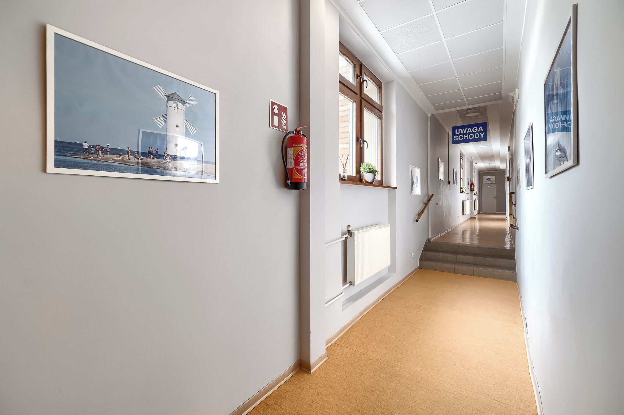 A hallway with a framed picture of a lighthouse, a fire extinguisher, and a sign reading "UWAGA SCHODY".