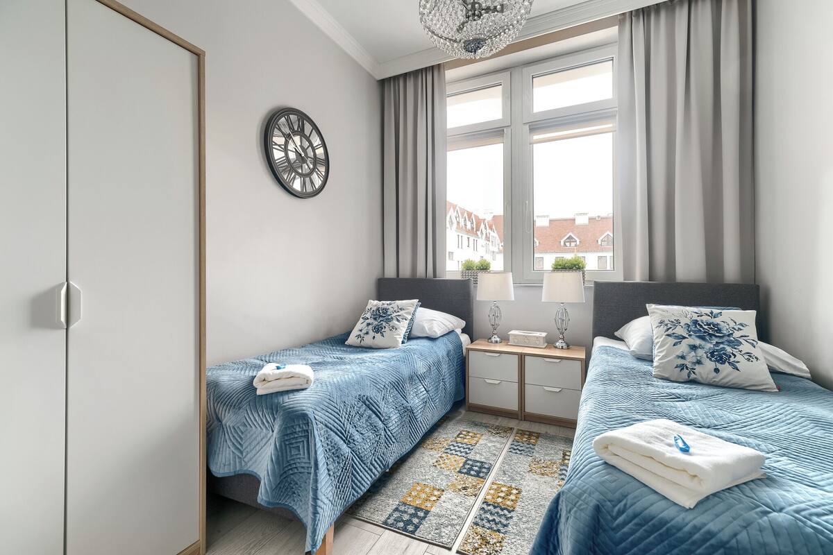 A bedroom with two beds, a large window, a clock on the wall, and a patterned rug.
