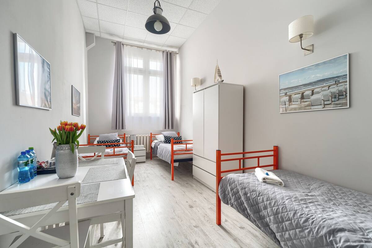 A modern, well-lit bedroom with a bed, a desk, and a chair. There are two bedside tables, a radiator, and a window with curtains.
