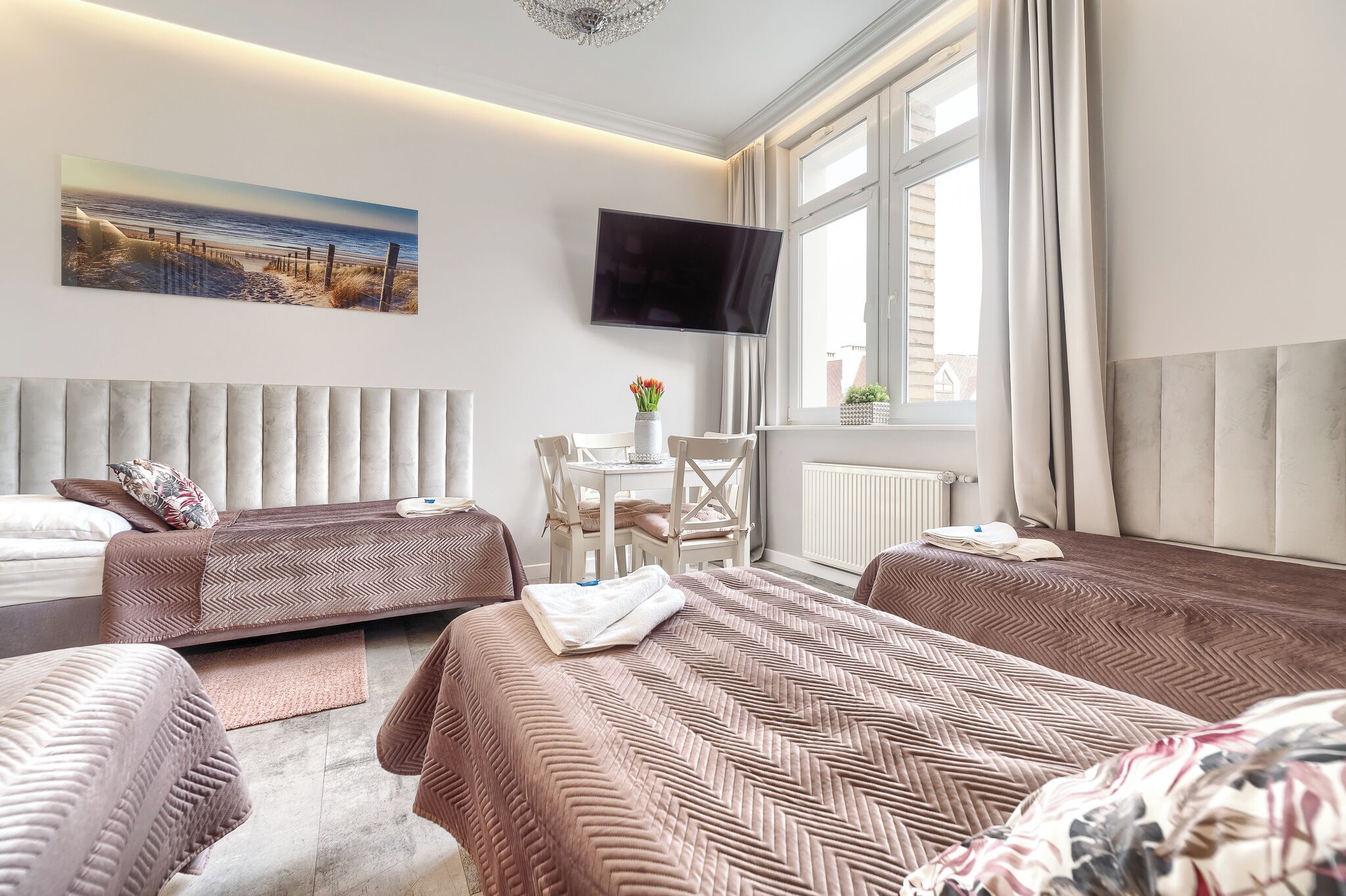 A hotel room with two beds, a TV, a window with curtains, a small table with a vase, and a painting on the wall.