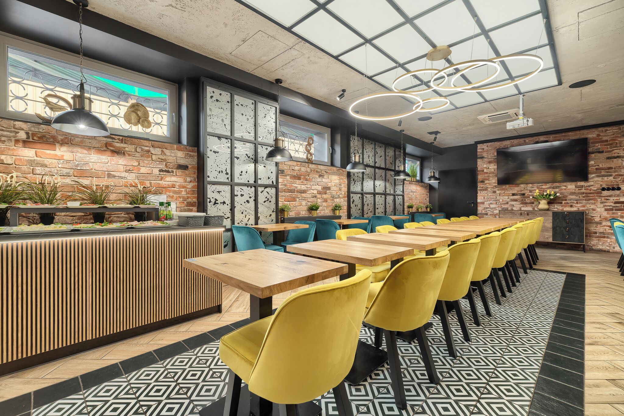 A modern restaurant with brick walls, wooden tables, yellow chairs, and a large TV screen.