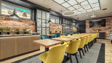 A modern restaurant with brick walls, wooden tables, yellow chairs, and a large TV screen.