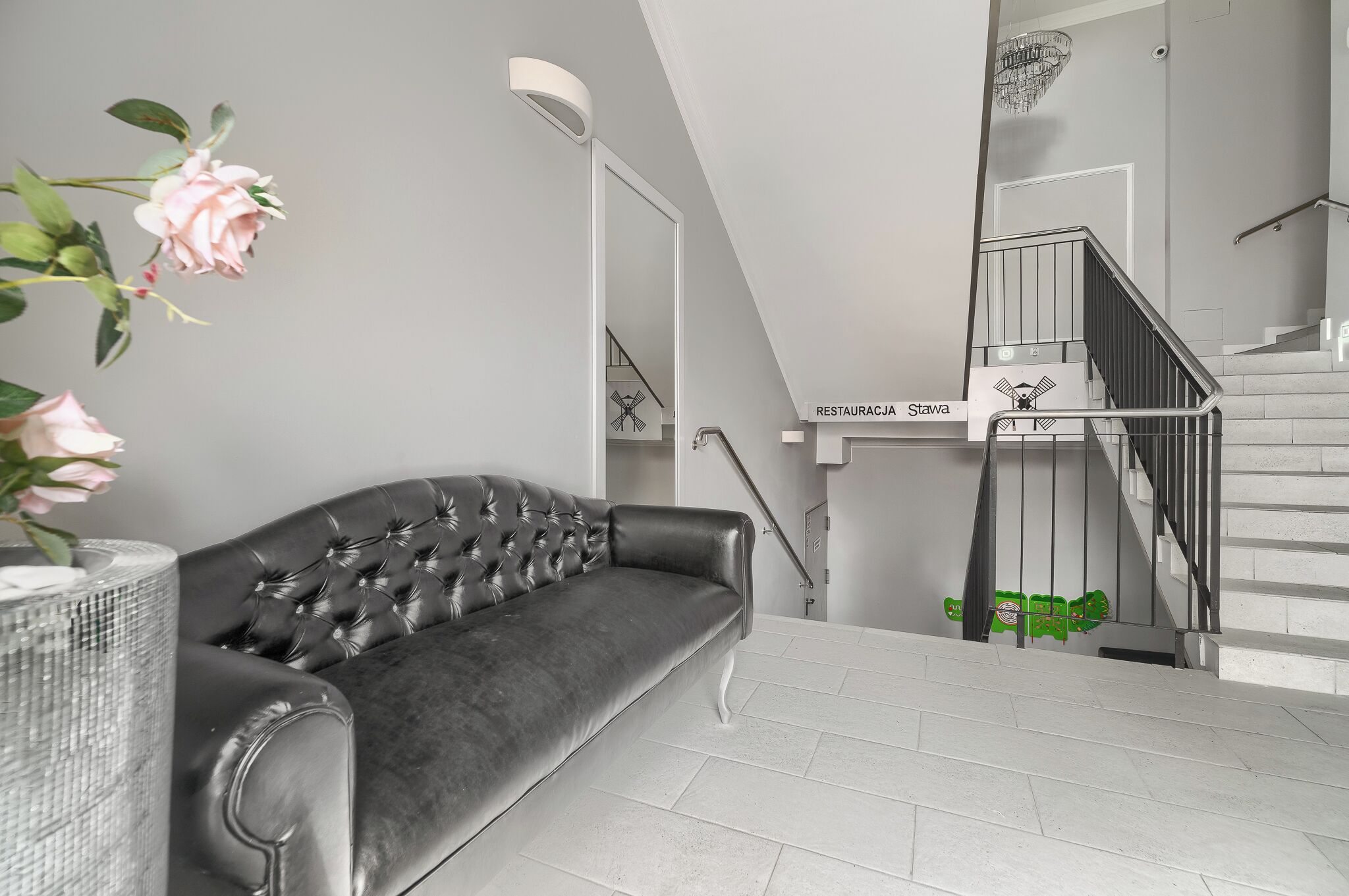 A modern living room with a dark leather sofa, a vase with pink flowers, and a staircase with a sign reading "RESTAURACJA Stała".