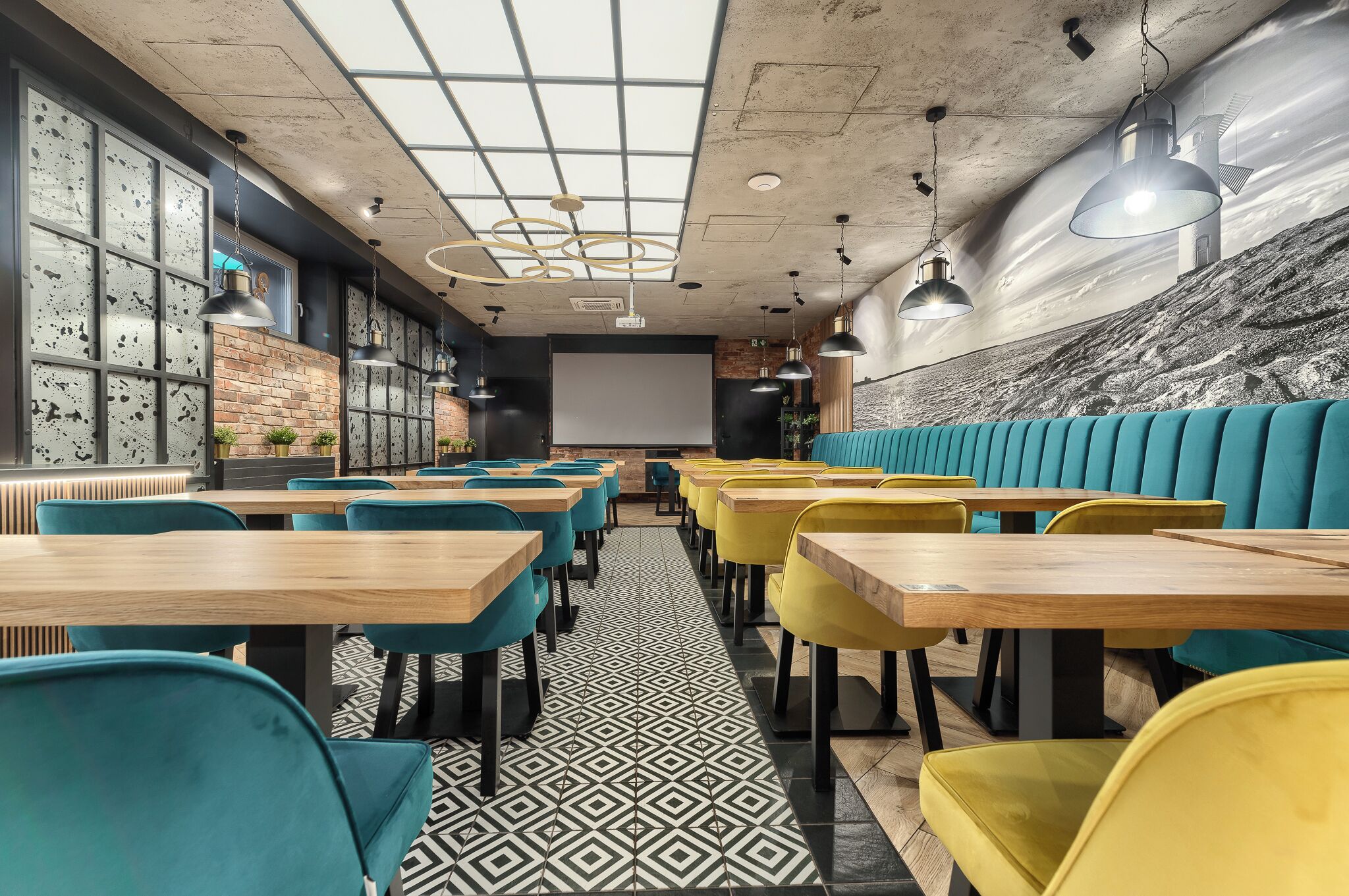 A modern restaurant with wooden tables and teal and yellow chairs, a patterned floor, and a large screen wall.