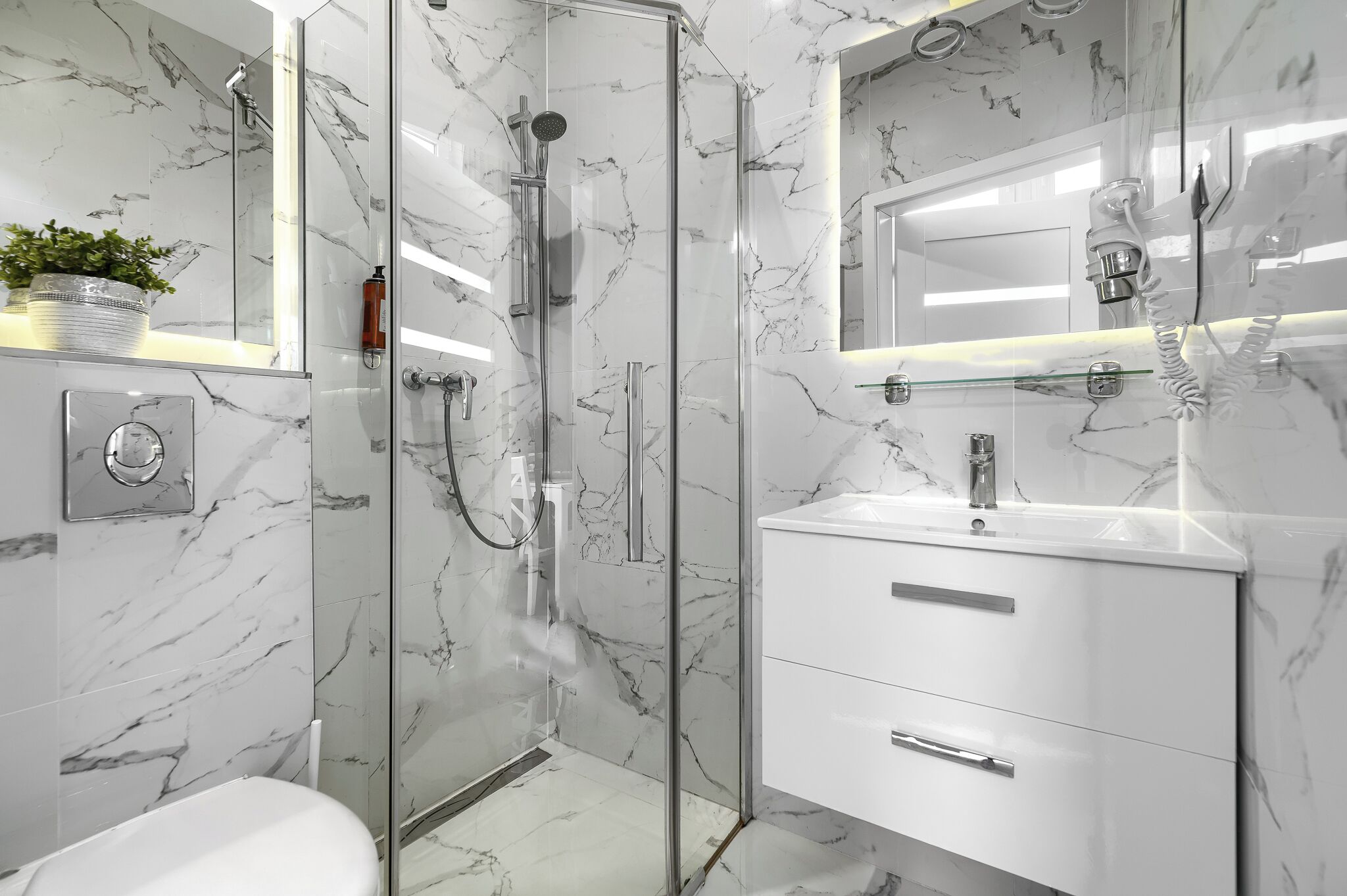 Un baño moderno con ducha de vidrio, paredes de mármol blanco y un mueble de tocador blanco con cajones.