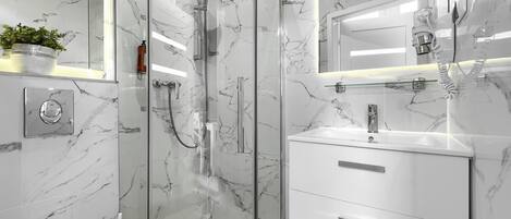 Un baño moderno con ducha de vidrio, paredes de mármol blanco y un mueble de tocador blanco con cajones.