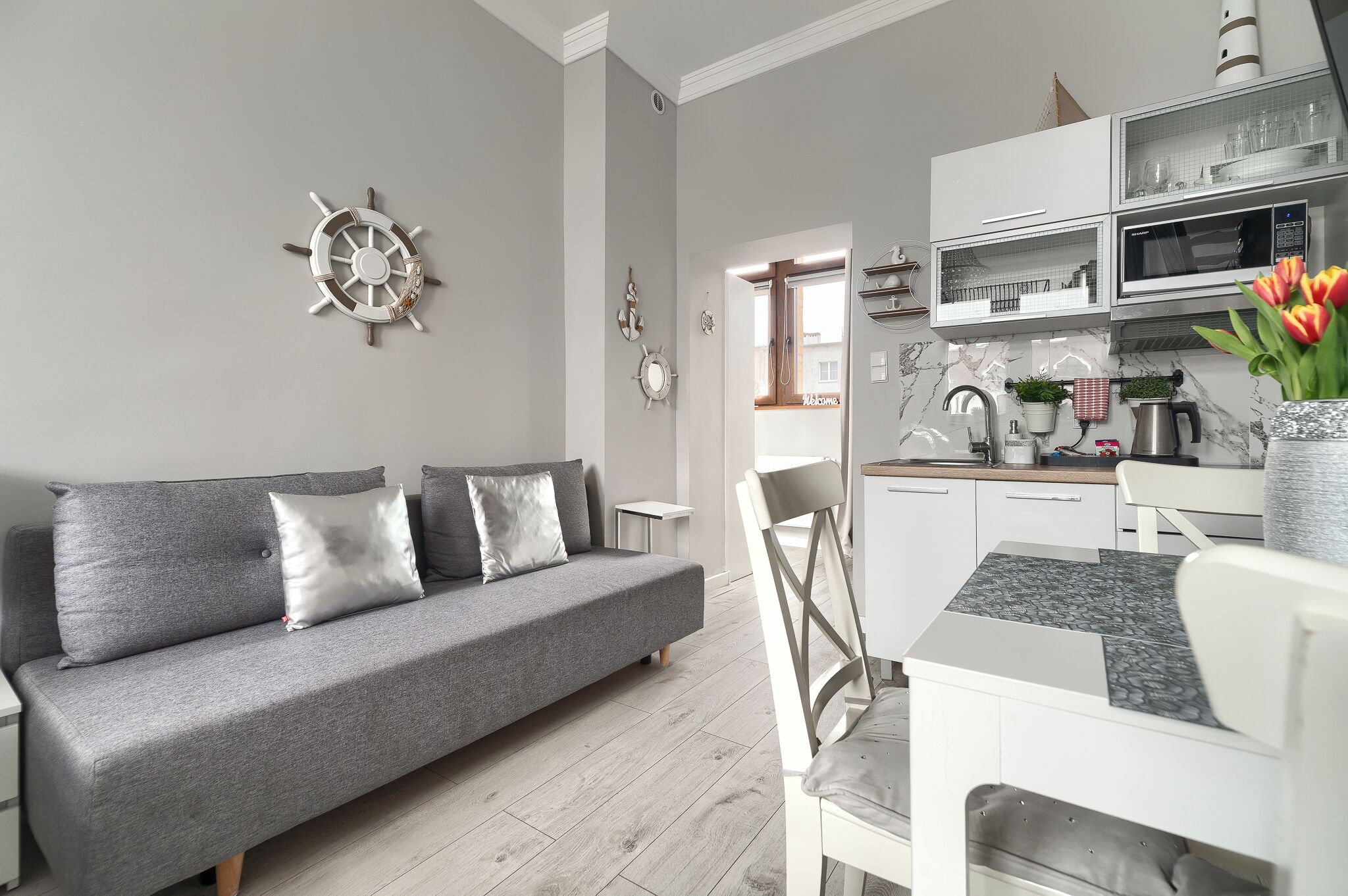 A modern living room with a grey sofa, a dining table with chairs, a kitchen area with white cabinets and a microwave, and a decorative ship's wheel on the wall.