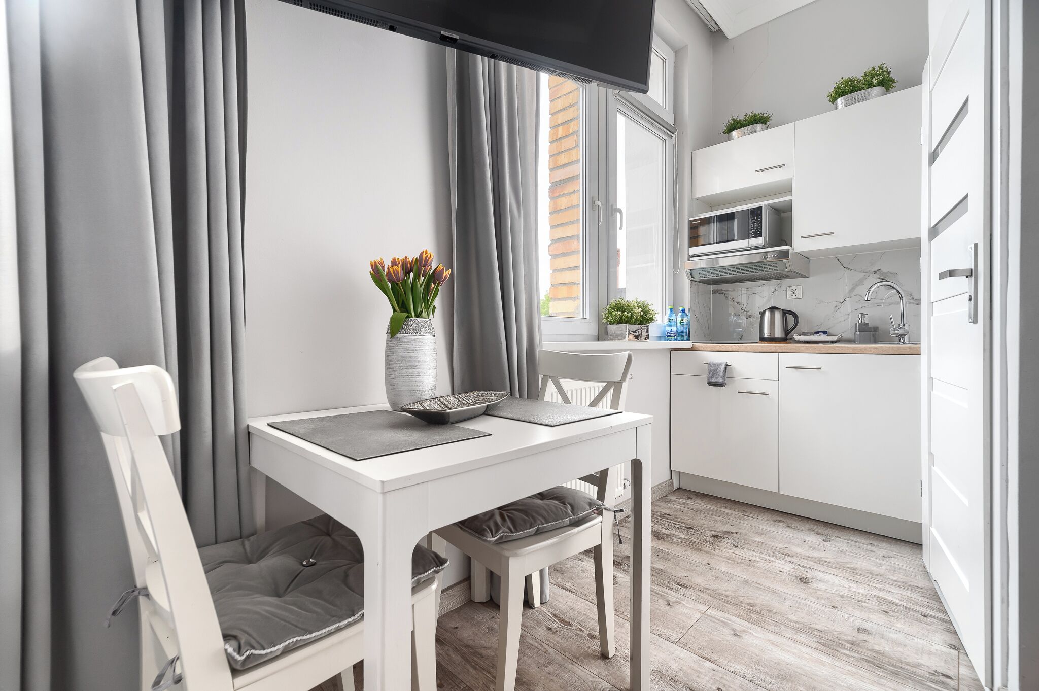 A modern kitchen with white cabinets, a microwave, and a kettle. There is a dining table with chairs and a vase of flowers.