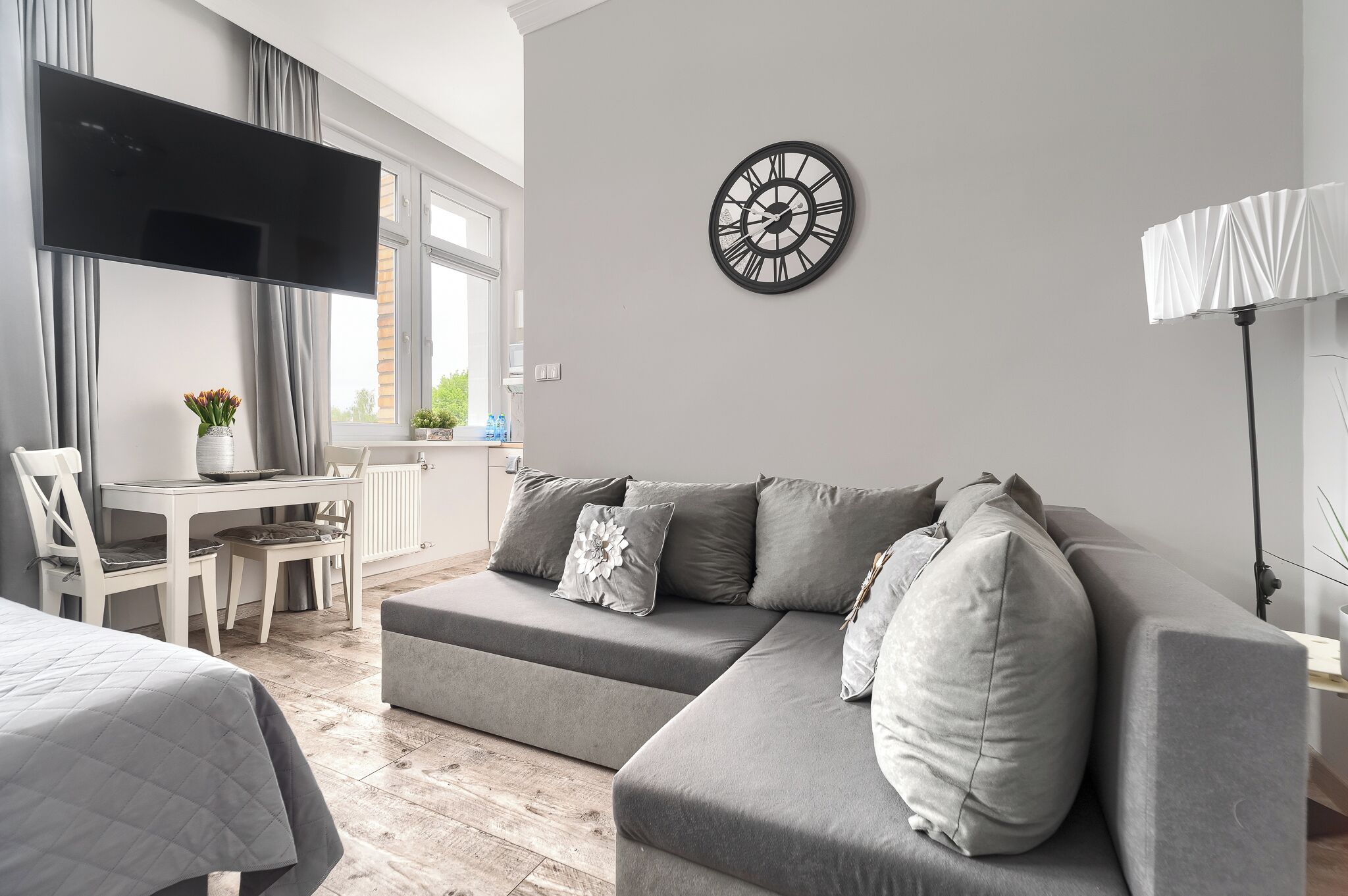 Una sala de estar moderna con un sofá gris, un televisor de pantalla plana, un reloj de pared redondo y un comedor con una mesa y sillas.