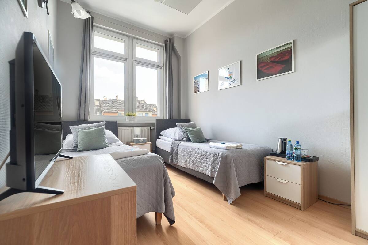A modern hotel room with two beds, a desk, a TV, and a window with a view of buildings.