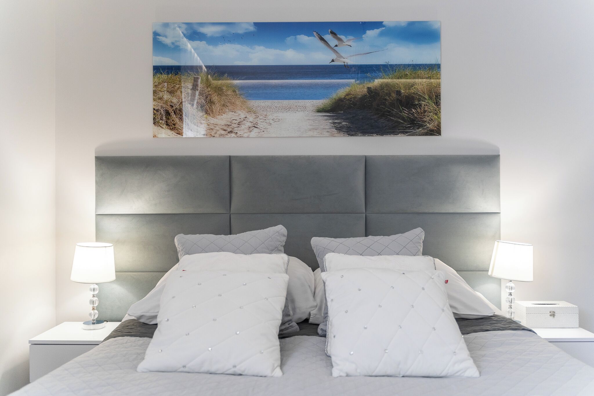 Une chambre à coucher avec un grand lit, deux tables de chevet, une tête de lit et un tableau représentant une scène de plage, accroché au mur.